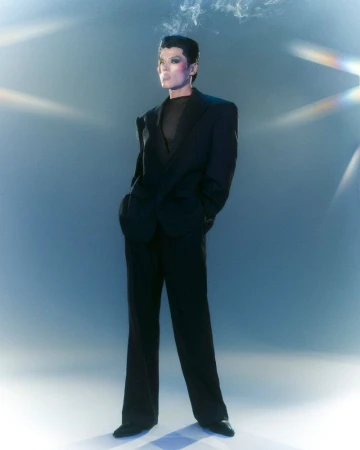 Stylish adult posing in a black suit against a blue background with smoke detail, standing in full shot in a fashion portrait.