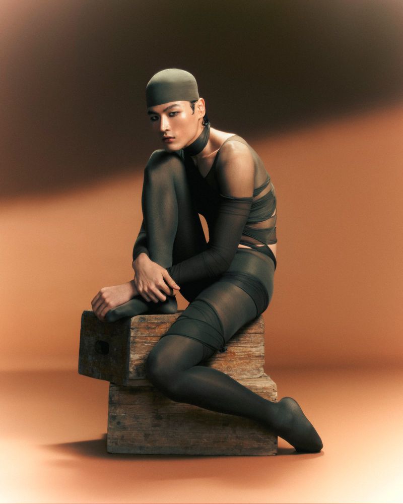 Male fashion model poses in a semi-sheer black top, tights, and head covering, seated on a rustic wooden block against a gradient backdrop.