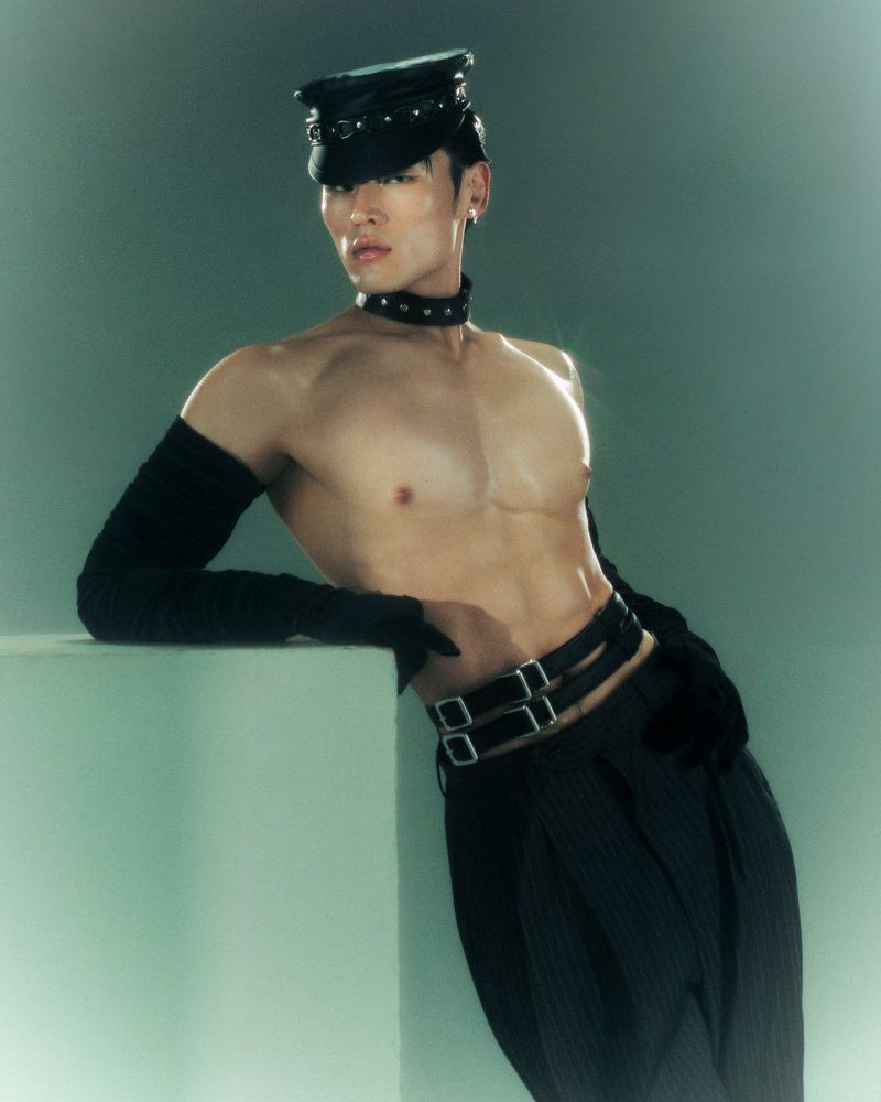 An Asian man strikes a bold pose in a photo studio, showcasing black leather accessories and pleated pants against an aquamarine backdrop.