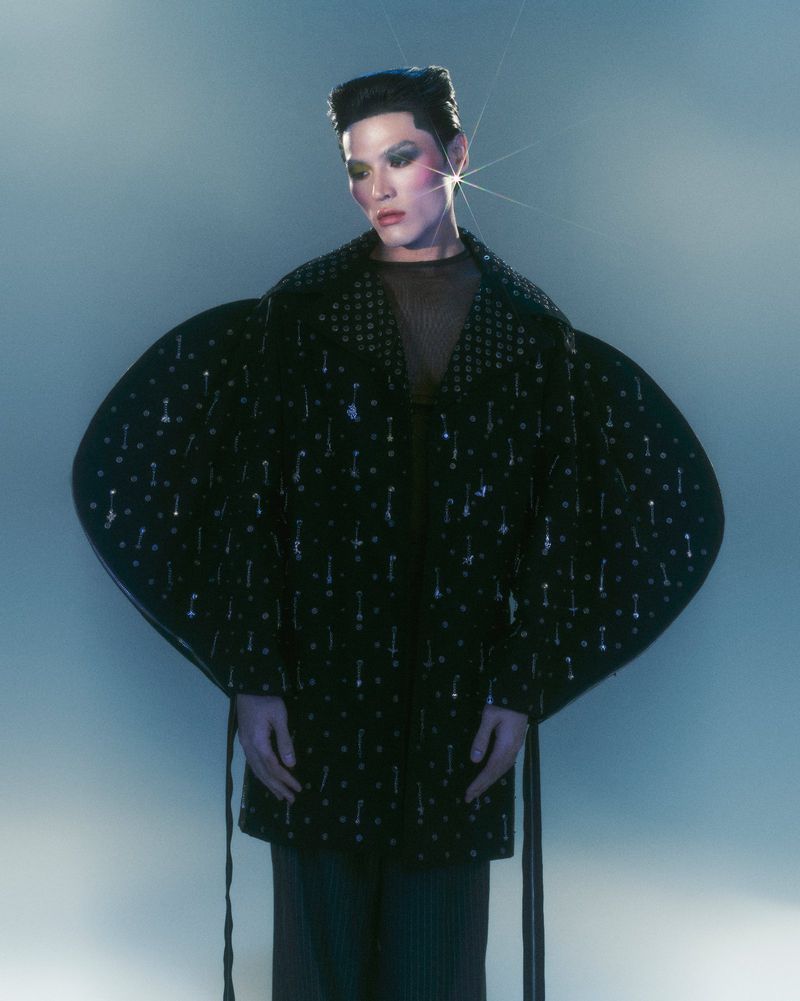 Model poses in dramatic light wearing black embellished coat with oversized sleeves and high-fashion makeup in an indoor setting.
