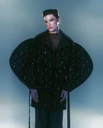 Model poses in dramatic light wearing black embellished coat with oversized sleeves and high-fashion makeup in an indoor setting.
