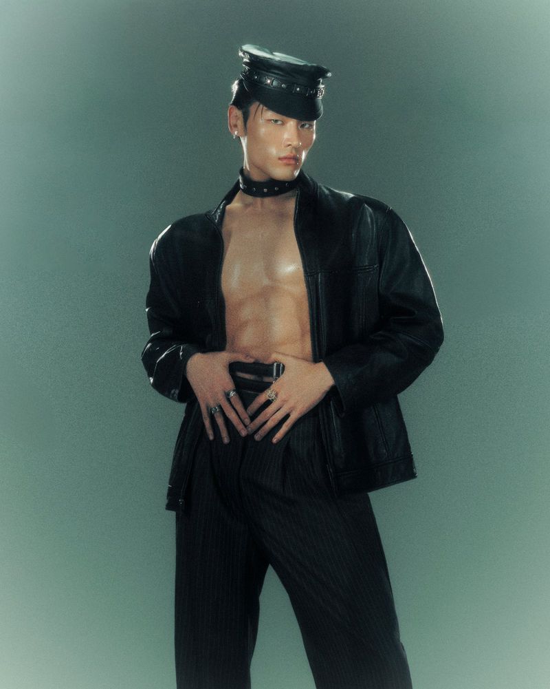 Stylish Asian man in leather jacket, choker, and cap poses confidently against a grey backdrop. Fashion editorial portrait.