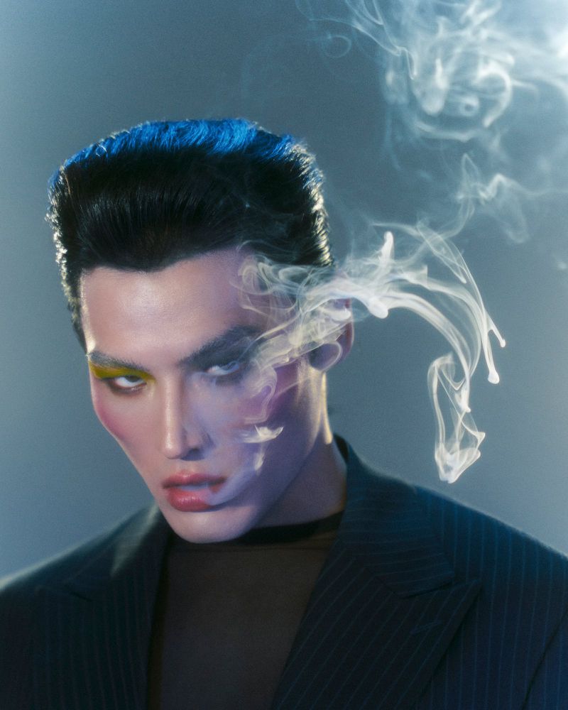 Dramatic portrait of a man with striking makeup and stylized hair exhaling smoke against a soft blue background in a studio setting.