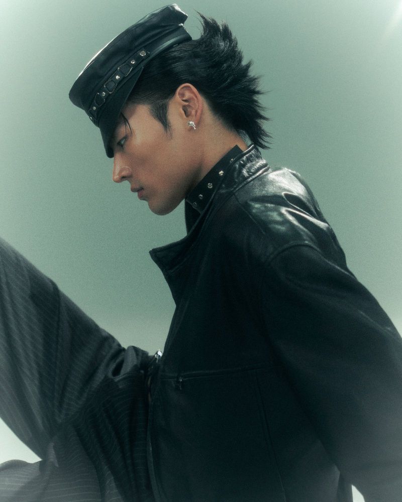 Profile shot of a person with black hair wearing a black leather jacket and studded cap against a light green studio backdrop.