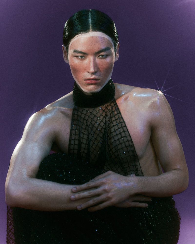 Close-up studio shot of a person with slicked-back hair, partially nude with black mesh fabric, and a purple backdrop with light flare.