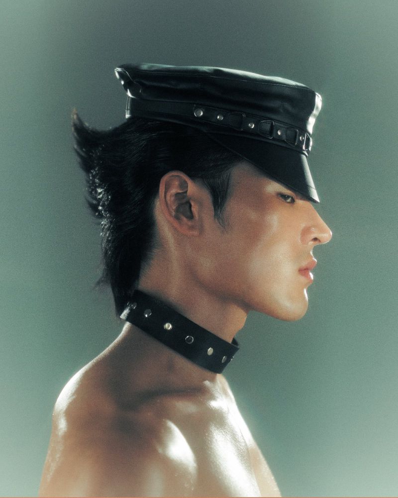 Profile portrait of a shirtless adult male wearing a black leather cap and collar with silver accents against a muted green background.