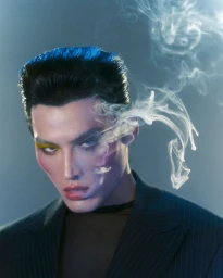 Portrait of a man with styled dark hair and yellow eye makeup wearing a dark suit jacket against a smoky blue background.