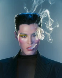 Portrait of a man with styled dark hair and smoke swirling around his face wearing a dark pinstripe blazer against a blue backdrop