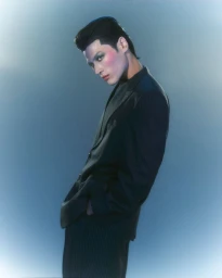 Model Craig Uy wearing a dark blazer with dramatic makeup against a gradient blue background for Fucking Young.