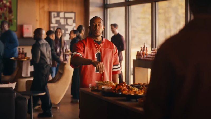 Man in a red jersey attends an indoor gathering with food, bottles, and people in the background in a well-lit room with large windows.