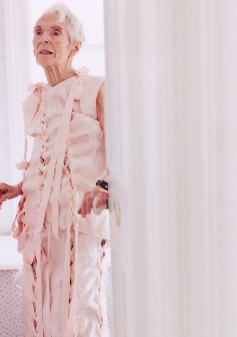 Elegant elderly woman in light pink lace-up attire stands beside a white wall, creating a timeless, soft-toned fashion portrait.