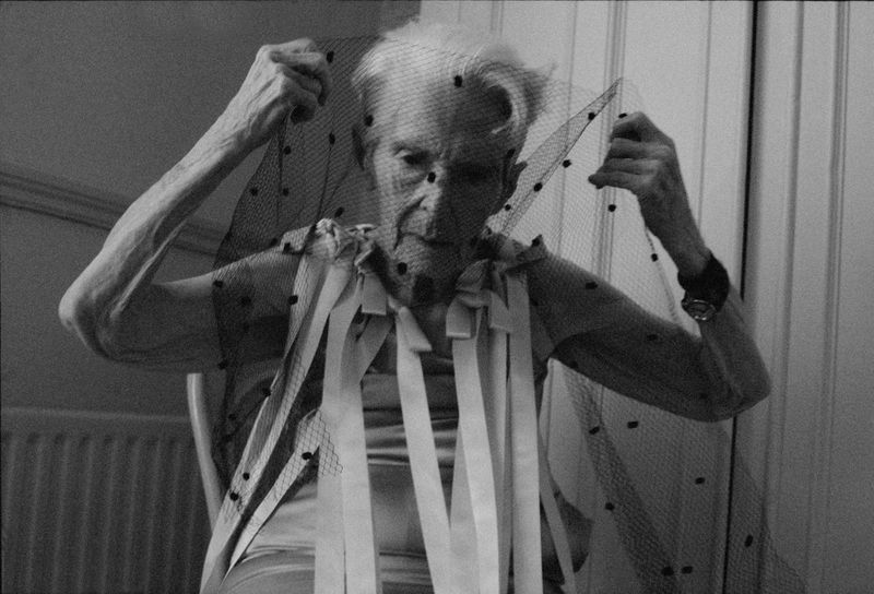 Elderly woman with short white hair poses in a black and white image, holding a mesh veil and wearing a striped top indoors.