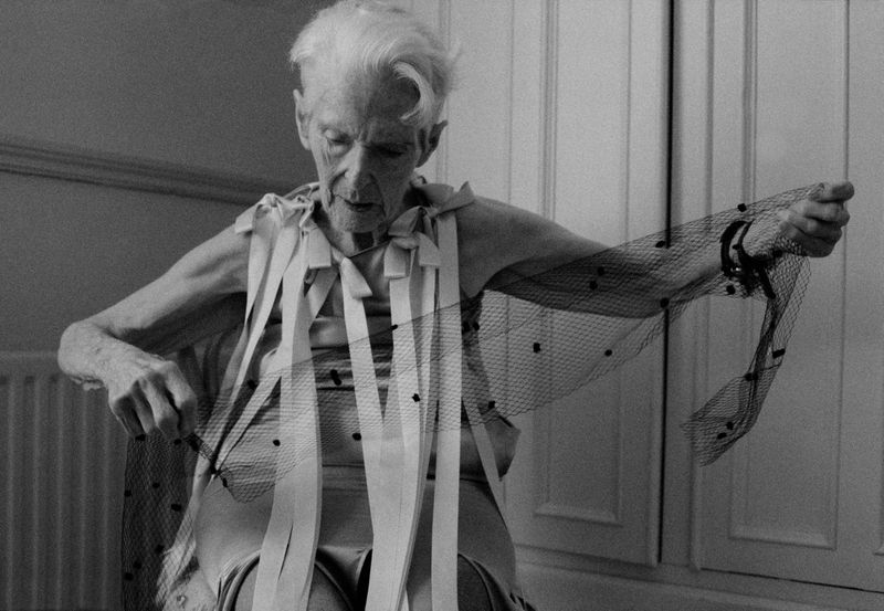Elderly adult in indoor setting poses with a black, dotted net fabric and white ribbon accents, in a monochrome shot.