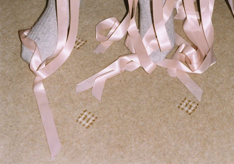 Feet in socks with pink satin ribbons tied around them on tan carpet with square pattern, highlighting texture and color contrast.