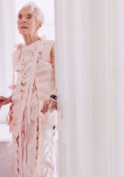Elderly woman in a light pink sleeveless top and trousers with ribbon ties standing in an indoor setting.