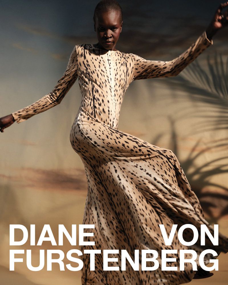 Fashion shot of a Black female model in a full-length cheetah print dress, posing outdoors with blurred natural background and brand text.