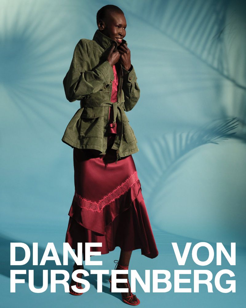 Model Alek Wek wearing an olive green jacket and burgundy skirt against a blue background with palm shadows for Diane von Furstenberg.