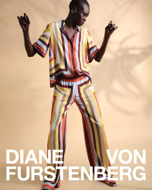 Model Alek Wek wearing a colorful striped silk shirt and matching trousers against a warm studio background.