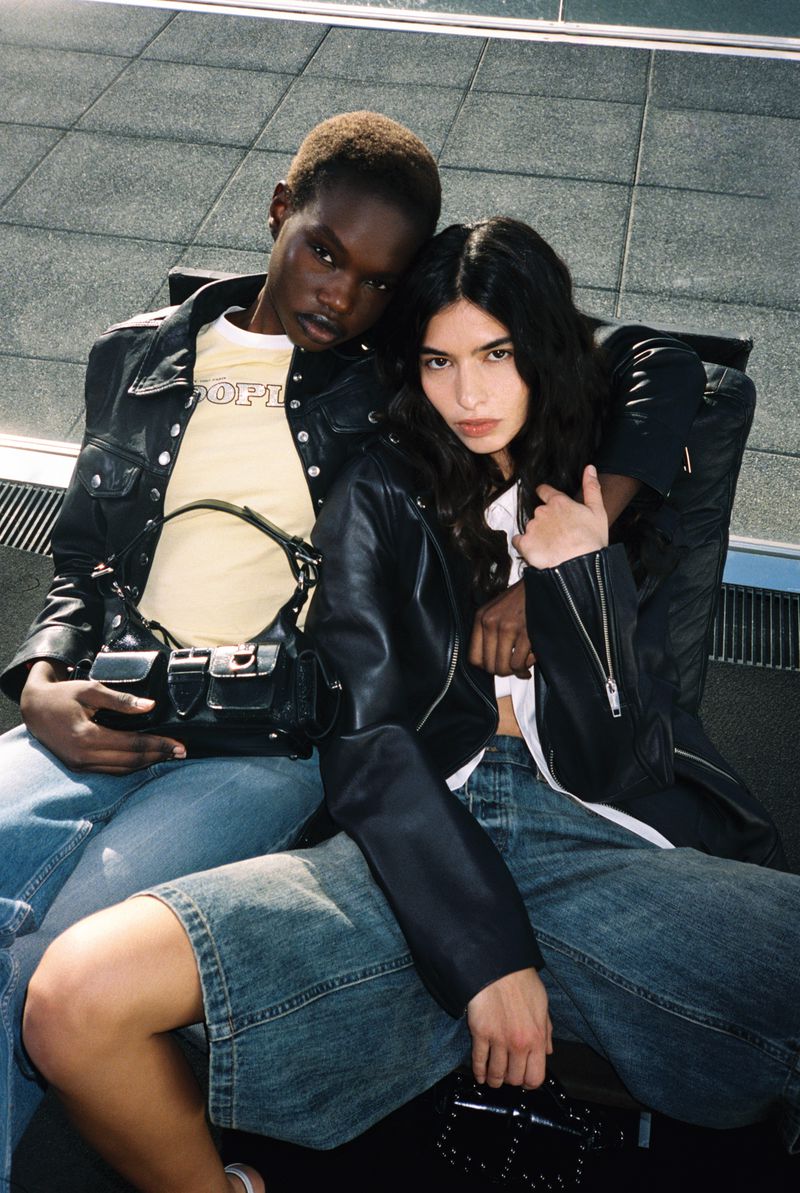 Two models pose together, wearing leather jackets and denim. One model wears a yellow tee and carries a black satchel bag with silver hardware.