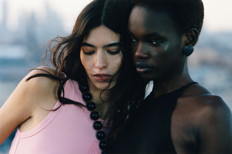 Two women in close-up view. One has tan skin and a pink top with a beaded necklace; the other has dark skin and a black top.