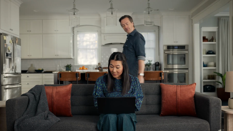 Woman using a laptop on a grey couch with a blanket and orange pillows in a modern home interior, with a man in the background.