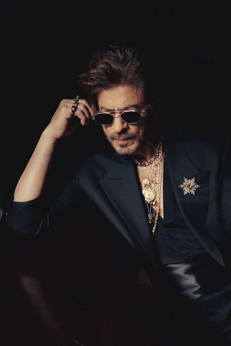 Portrait of a stylish adult male with sunglasses wearing a dark suit jacket and multiple gold necklaces against a dark background.
