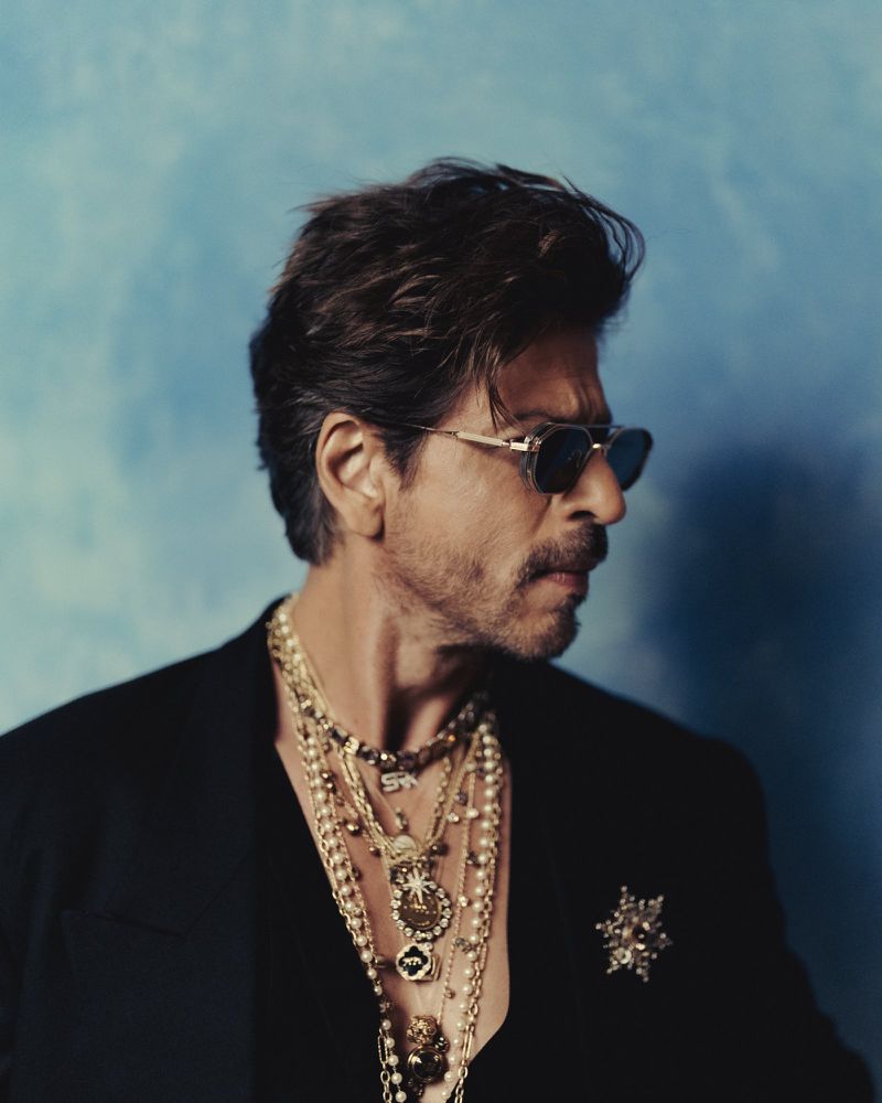 Close-up portrait of a mature adult male wearing sunglasses and multiple gold necklaces with a suit jacket against a textured blue background.