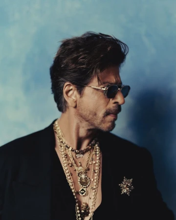Close-up portrait of a mature adult male wearing sunglasses and multiple gold necklaces with a suit jacket against a textured blue background.