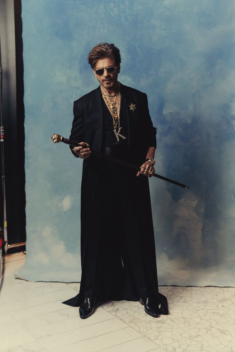 Adult male in a black suit and sunglasses holds a cane, standing in a photo studio against a blue backdrop, with a confident expression.