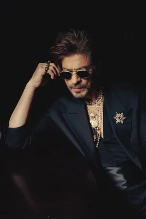 Shah Rukh Khan wearing a dark suit, sunglasses, and layered gold chains in a low-key portrait.