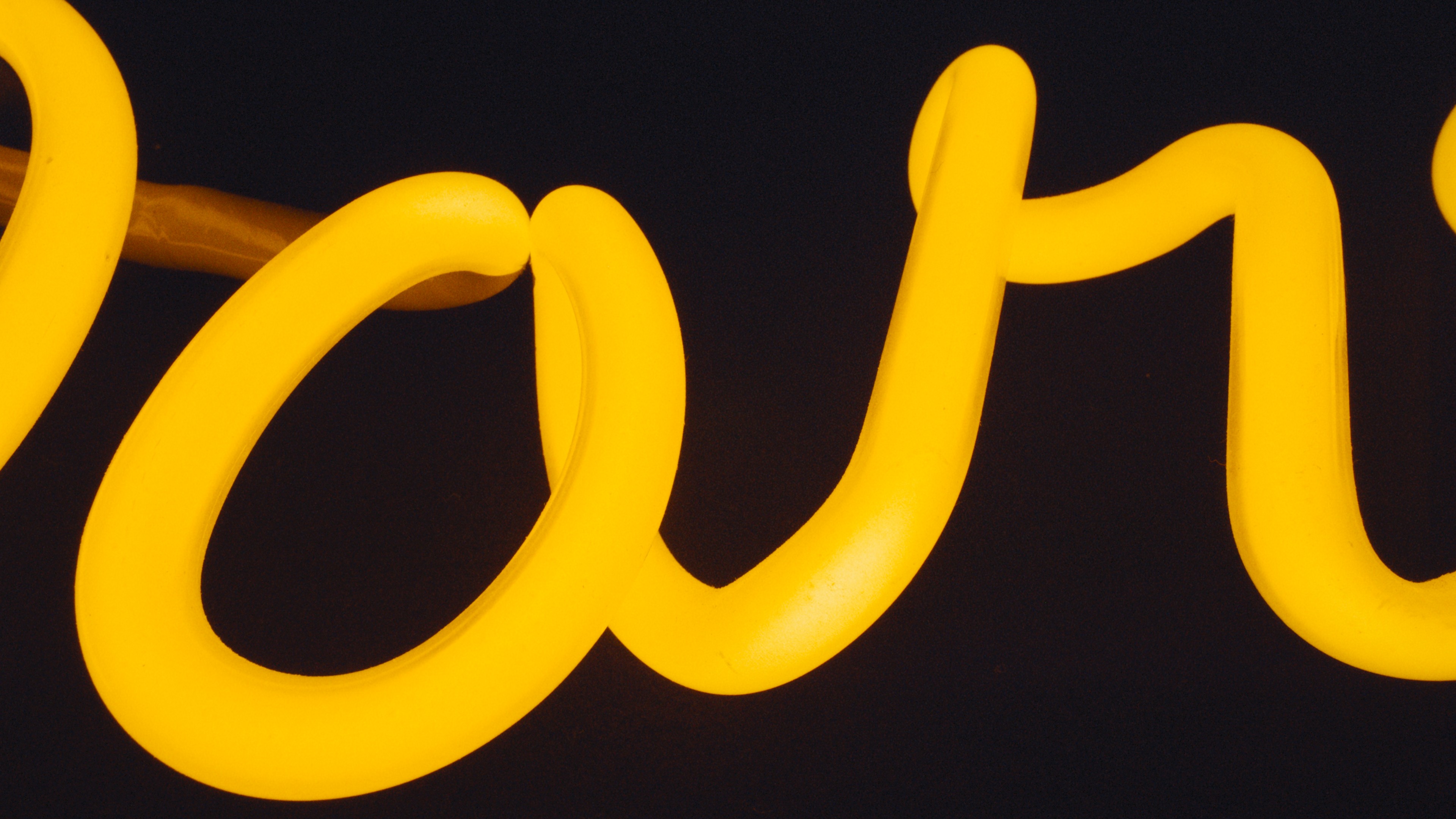 Close-up studio shot of yellow neon cursive text against a dark background with a blurry brown support rod visible.