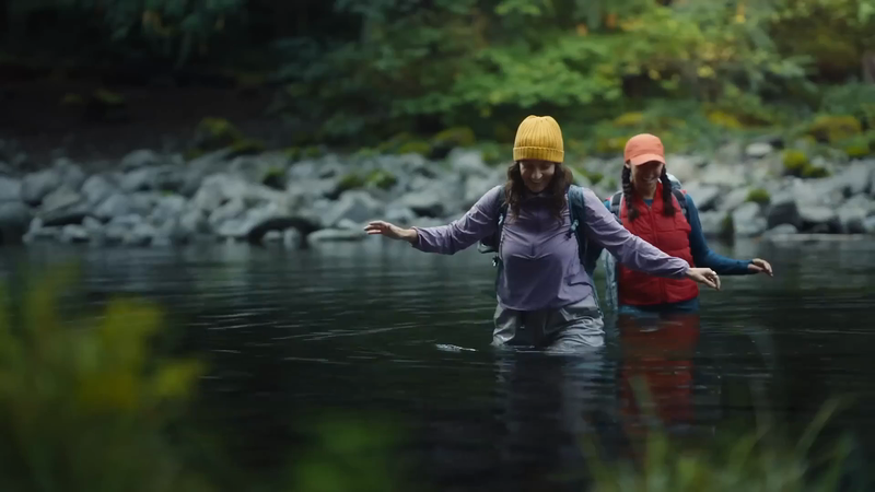 Two adult females are crossing a shallow river. They wear hats and backpacks. The water is dark, with rocks and green trees in the background.
