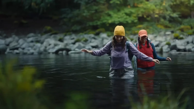 Two adult females are crossing a shallow river. They wear hats and backpacks. The water is dark, with rocks and green trees in the background.