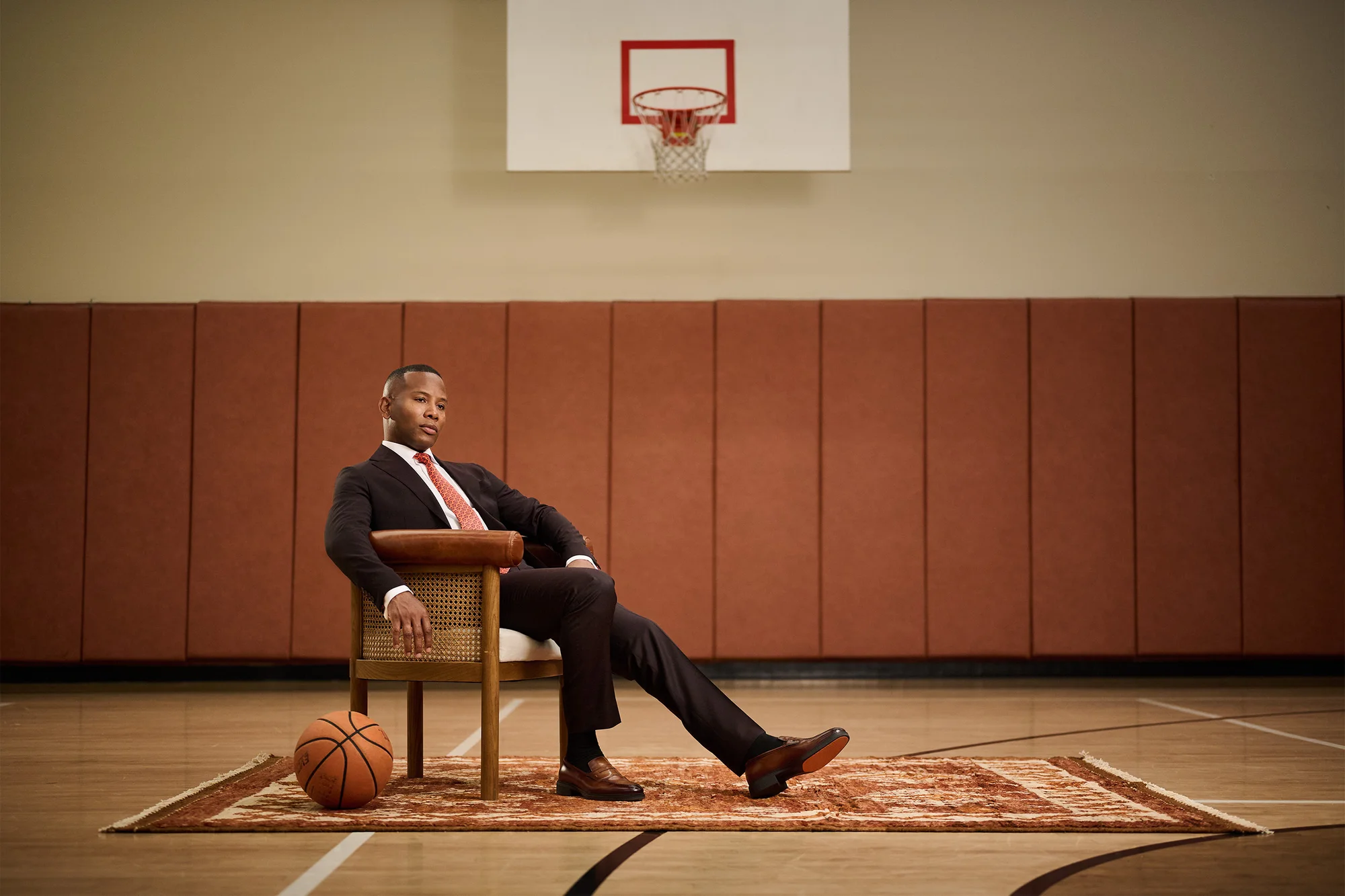 Man in suit sits on chair in basketball court with ball and rug. Indoor portrait, athletic setting, professional attire, stylish.