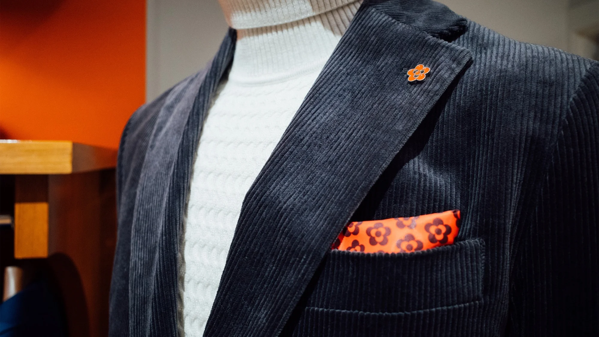 A grey corduroy blazer is styled with a white turtleneck sweater, accented by an orange pocket square and flower lapel pin.