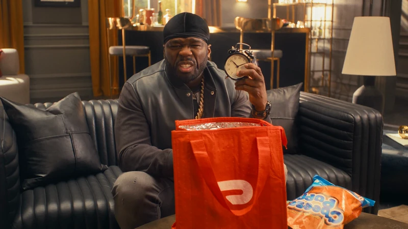 50 Cent poses indoors holding an alarm clock next to a DoorDash delivery bag, wearing casual clothes in a living room setting.