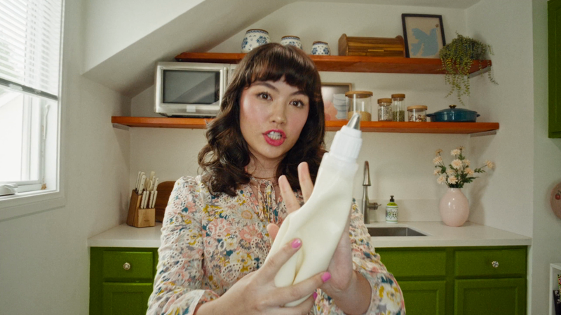Woman holds piping bag in kitchen setting with green cabinets and natural light, demonstrating culinary skills for online cooking content.