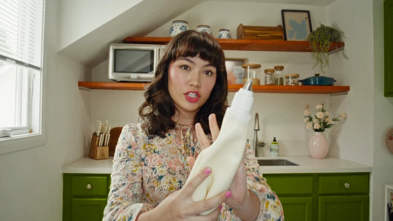 Woman holds piping bag in kitchen setting with green cabinets and natural light, demonstrating culinary skills for online cooking content.