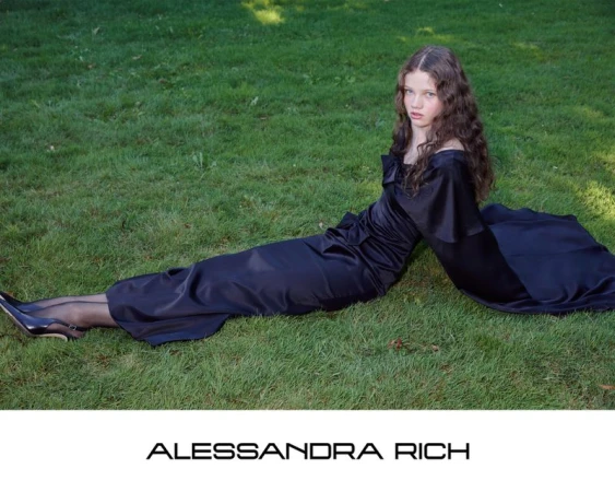 Young woman with long dark wavy hair wearing a black satin evening dress reclining on a green grass lawn.