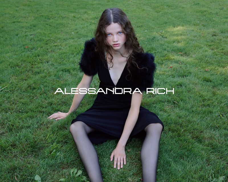 Young woman in a black dress with fur shoulder accents poses seated on green grass in a fashion editorial image by Alessanda Rich.