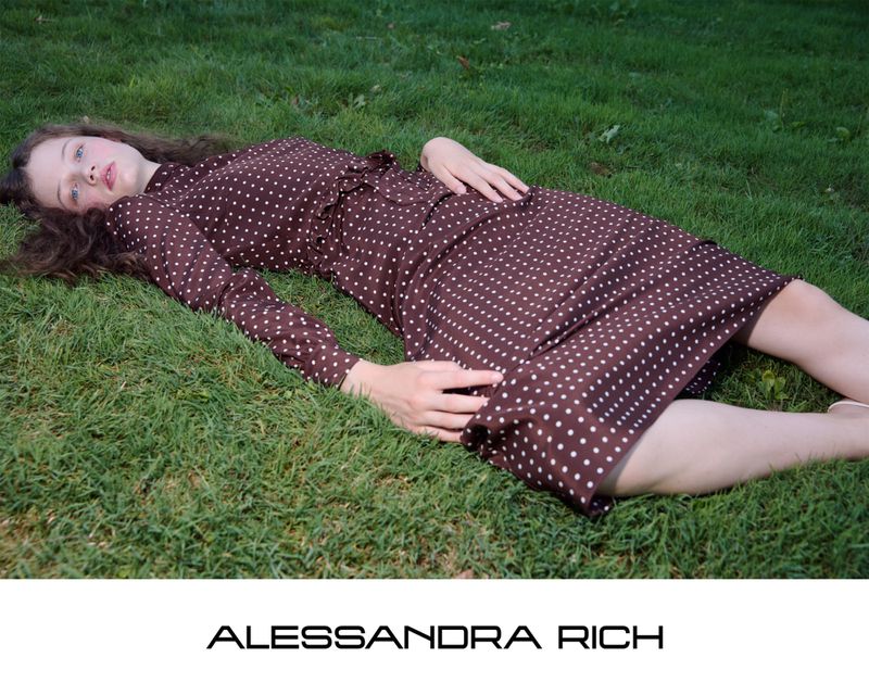 Model lying on green grass wearing a brown polka dot dress for the Alessandra Rich S/S 2026 campaign.