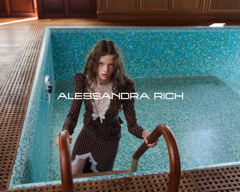 A young woman in a brown polka-dot dress stands on pool steps, holding the railing, in an indoor pool setting with natural light.