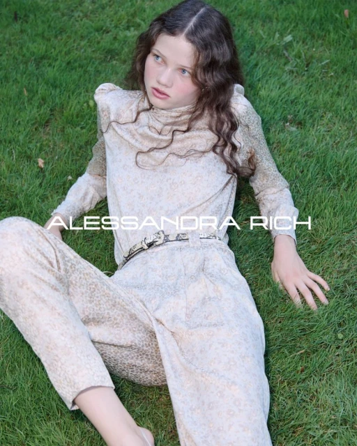 A girl wearing a patterned beige pantsuit lies on the grass outdoors, styled with a belt, and the brand name ALESSANDRA RICH is overlayed.