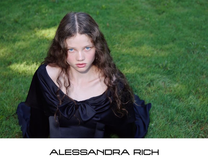 A young woman with long dark wavy hair wearing a black dress sits on green grass in the Alessandra Rich S/S 2026 campaign.