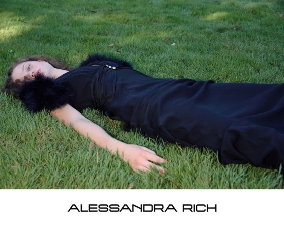 A woman wearing a black dress with feather trim lying on green grass for the Alessandra Rich S/S 2026 campaign.