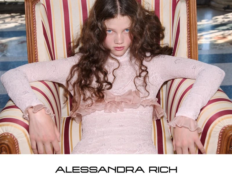 A young woman with curly brown hair wears a pink lace dress with ruffle detail while sitting in a red and tan striped armchair.