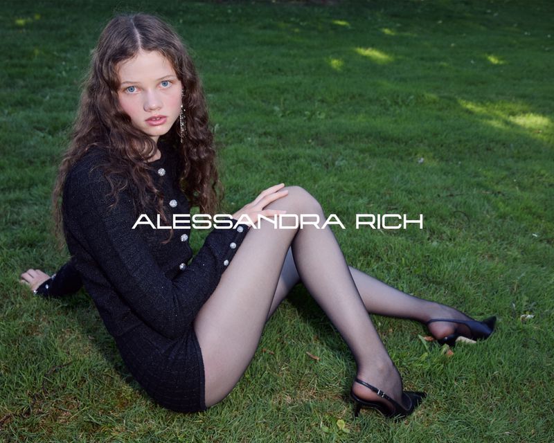 A young woman in a black dress and pantyhose poses on grass outdoors, with white text overlaying the image. Fashion editorial.