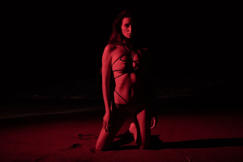 Adult female in bikini kneeling on sand, posing in a photo studio under dramatic red lighting against a dark backdrop.