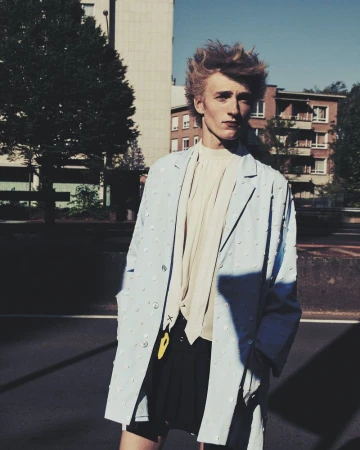 Young man in a light blue jacket and black shorts standing on an urban street with buildings and trees in the background on a sunny day.
