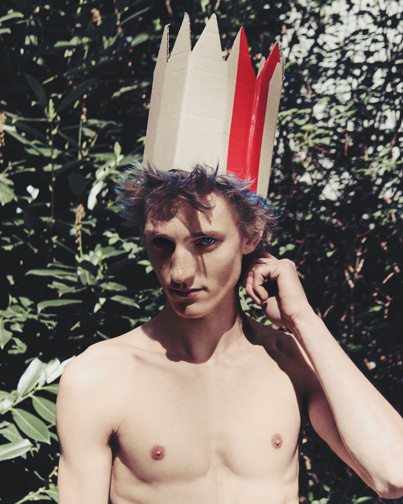 Portrait of a shirtless male with blue hair, wearing a cardboard crown with red accents, standing outdoors surrounded by green foliage.